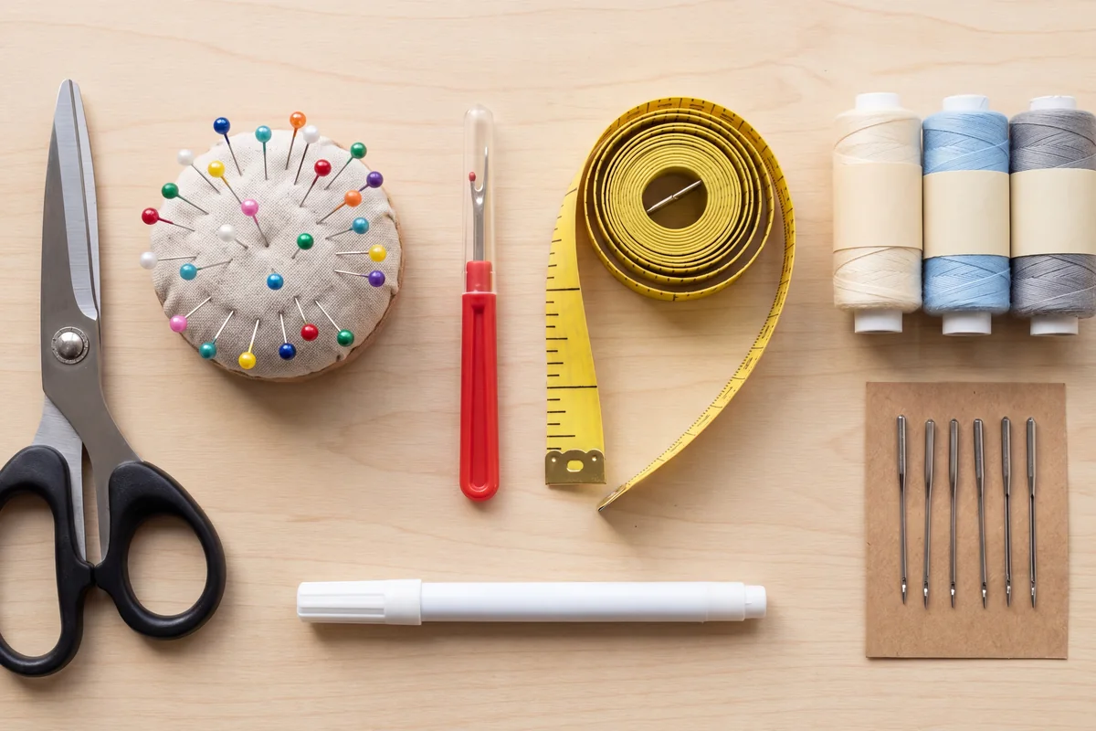 Beginner sewing kit on a wooden table with fabric shears, pins, seam ripper, tape measure, chalk marker, needles and thread spools