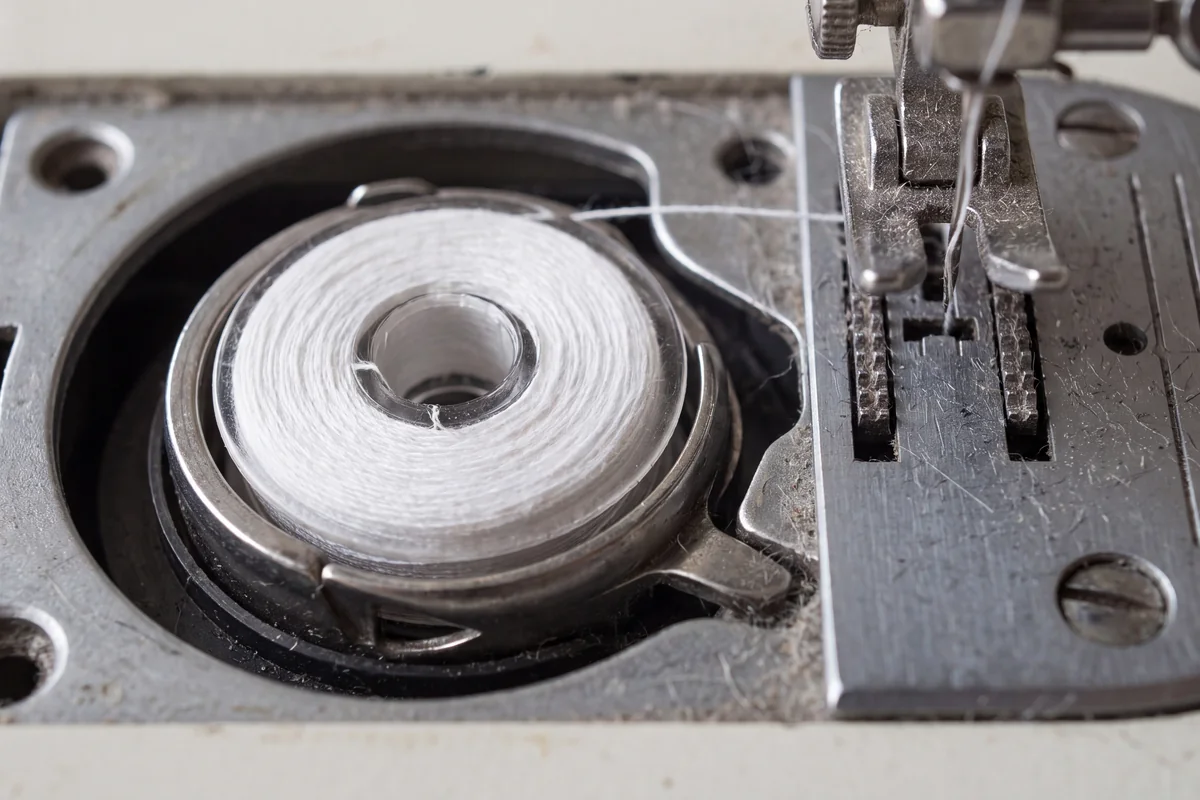 Drop-in bobbin with white thread in its holder next to the feed dogs on a sewing machine needle plate