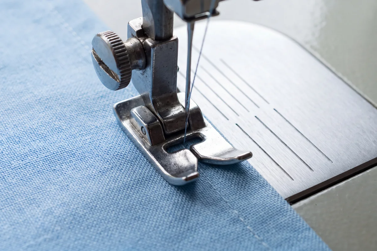 Sewing machine needle, presser foot and needle plate with engraved seam guide lines beside light blue cotton fabric