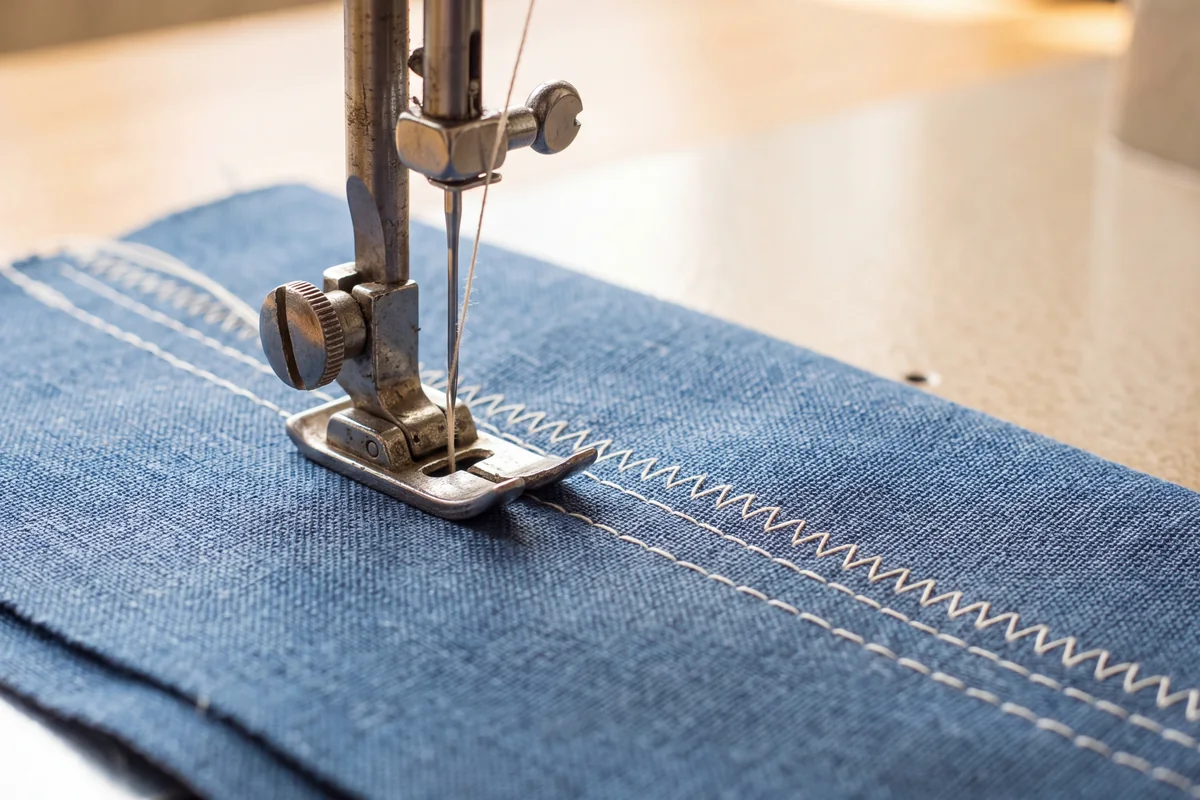 Sewing machine presser foot and needle on blue cotton next to a row of straight stitches and a row of zigzag stitches